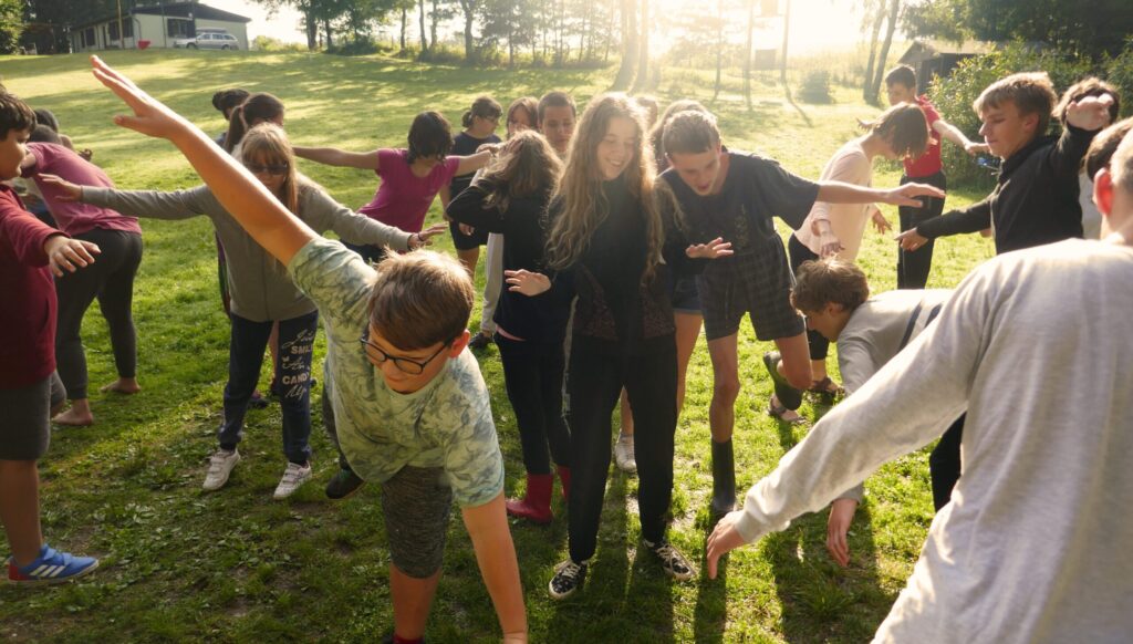 Na fotce je táborová parta. Je to momentka, kdy všichni dělají "letadlo", tedy mají rozpažené ruce a "letí". Někteří se u toho smějí, někteří dokonce dělají holubičku a stojí u toho na jedné noze. Celá fotka působí velmi pohodovým dojmem, za celou skupinou navíc prosvítají paprsky slunce.